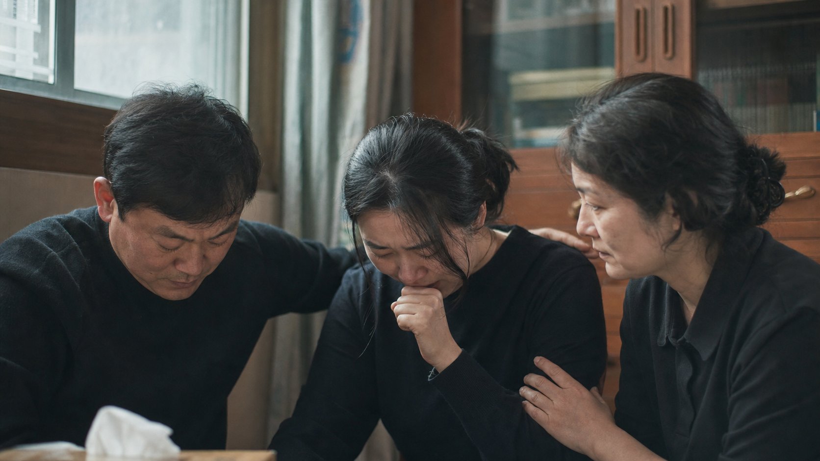 창가에서 가족이 슬픔에 잠긴 한 사람의 어깨를 감싸 안으며 함께 위로하는 따뜻한 장면
